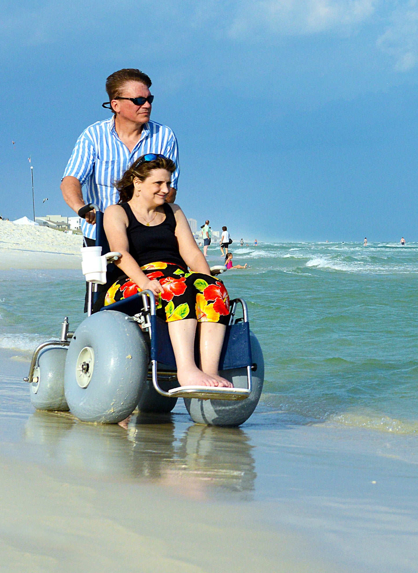 fixed frame beach wheelchair on the edge of the ocean in the sand with a woman in the chair and a man pushing her.