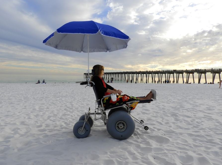 elevating leg rest beach wheelchair with a women sitting with her legs elevated and under an umbrella. They are in the sand at the beach.