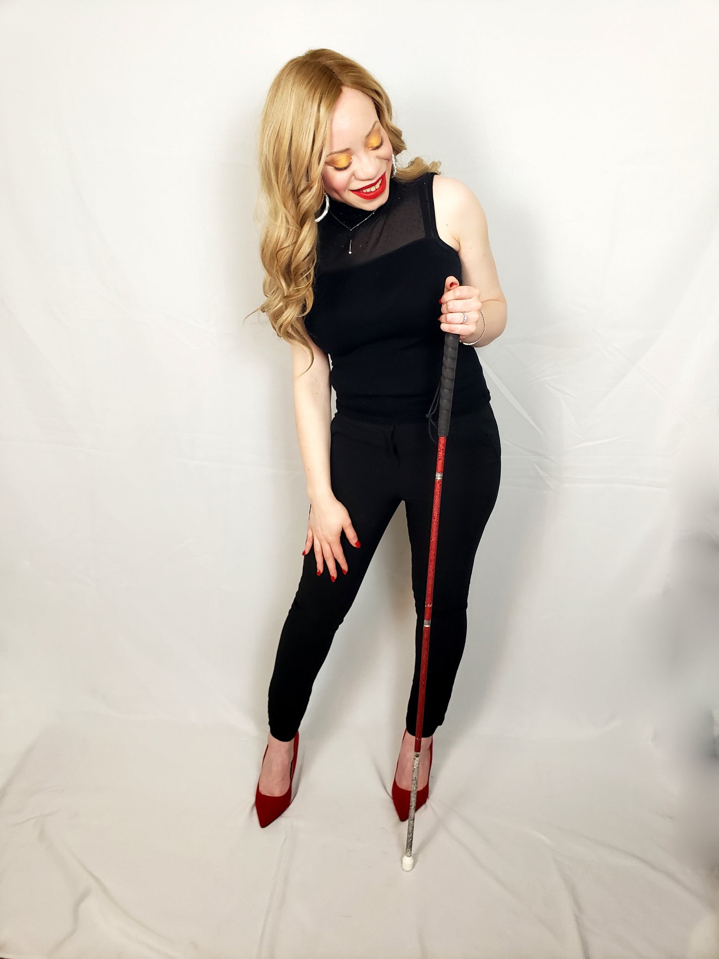 A woman poses confidently holding a rhinestone glam cane with a custom red upper 2/3 of the cane and white lower 1/3 of the cane design to match her red lipstick, nails, and red shoes.