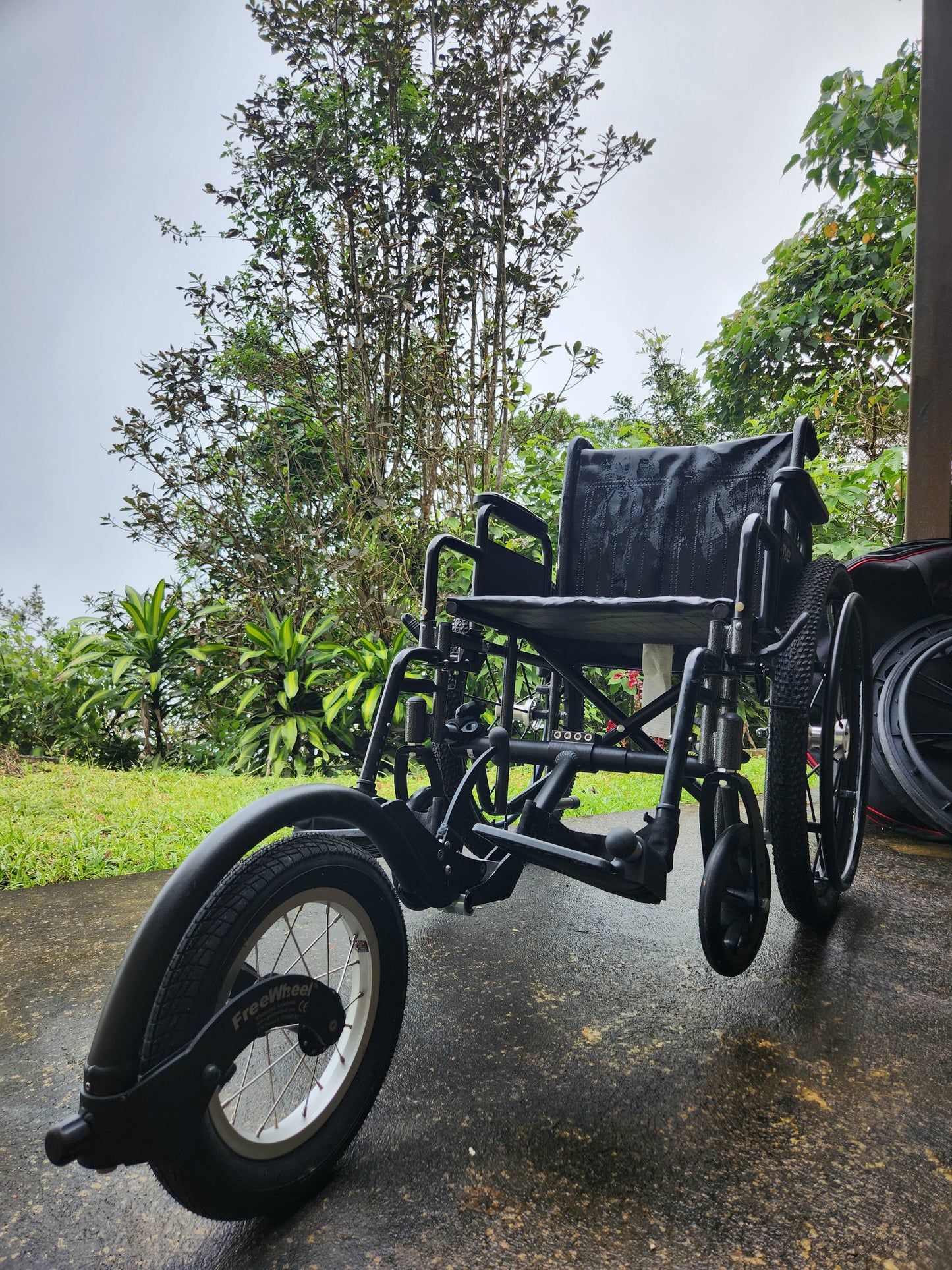 Black wheelchair with a 5th wheel on a paved road with trees in the background.