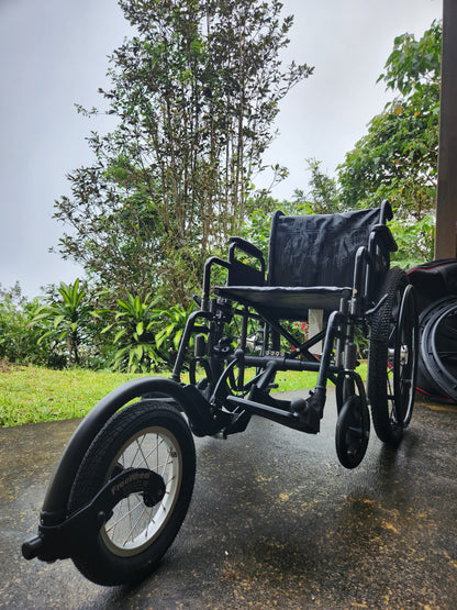 Black wheelchair with a 5th wheel on a paved road with trees in the background.