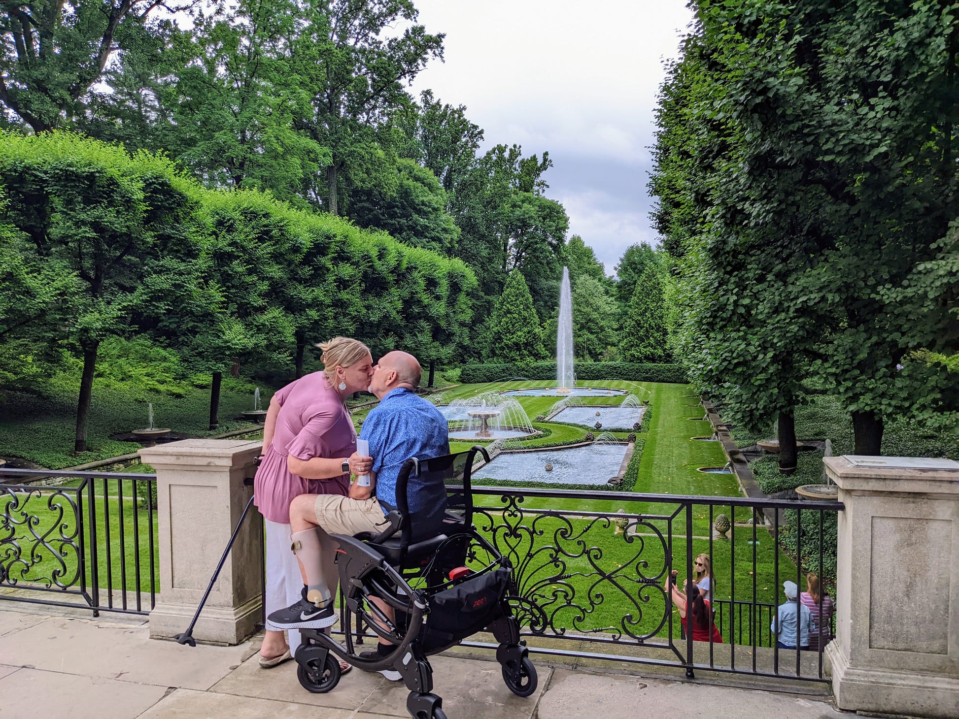 Two people, one in a wheelchair, sitting on a stone ledge overlooking a garden with a fountain.